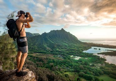 Young shirtless male with a backpack standing on a mountain and taking a picture under a cloudy sky